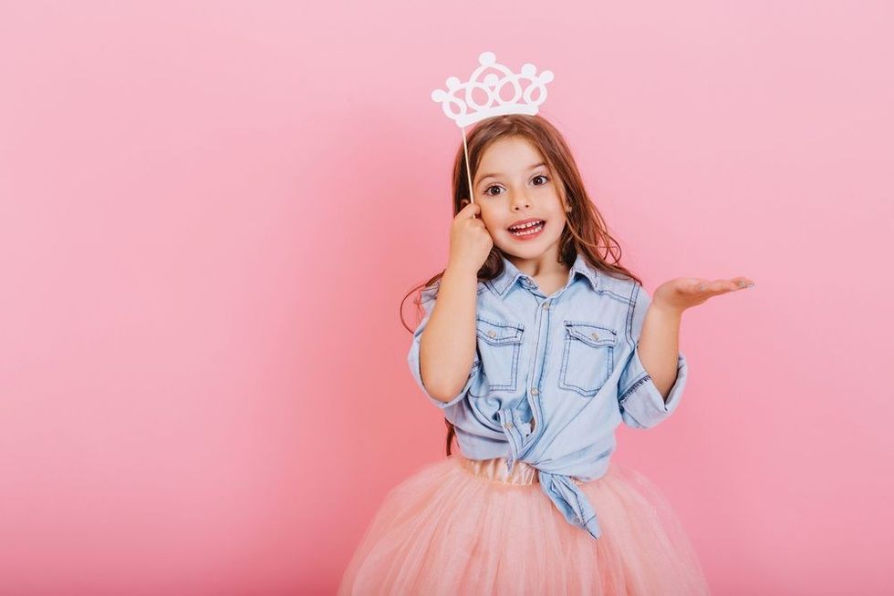 Joyful little girl with long brunette hair in tulle skirt holding princess crown on head.