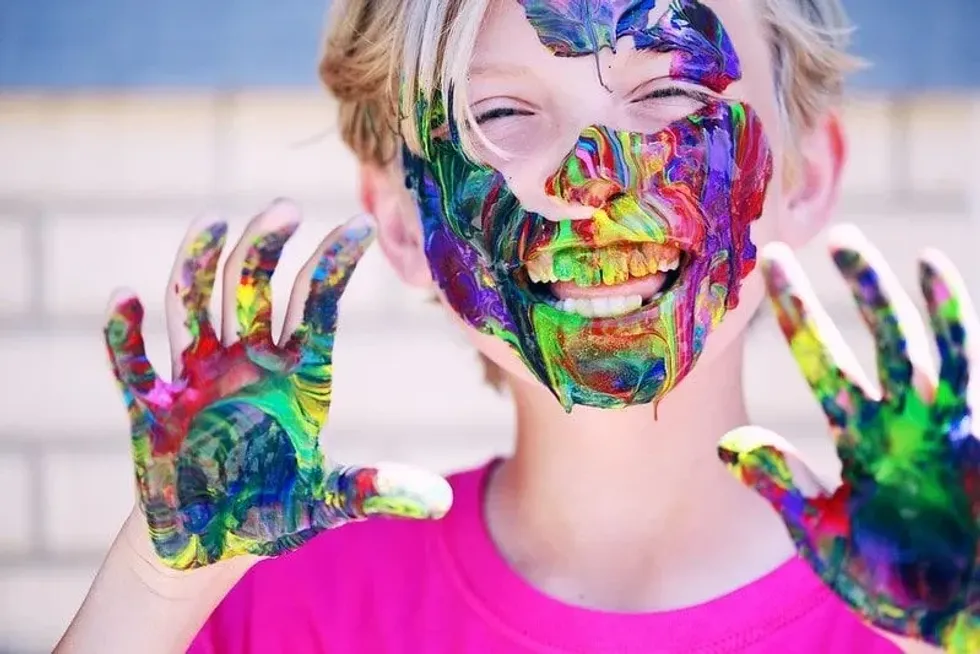 Kid smiling with colourful paint all over their hands, face and teeth.