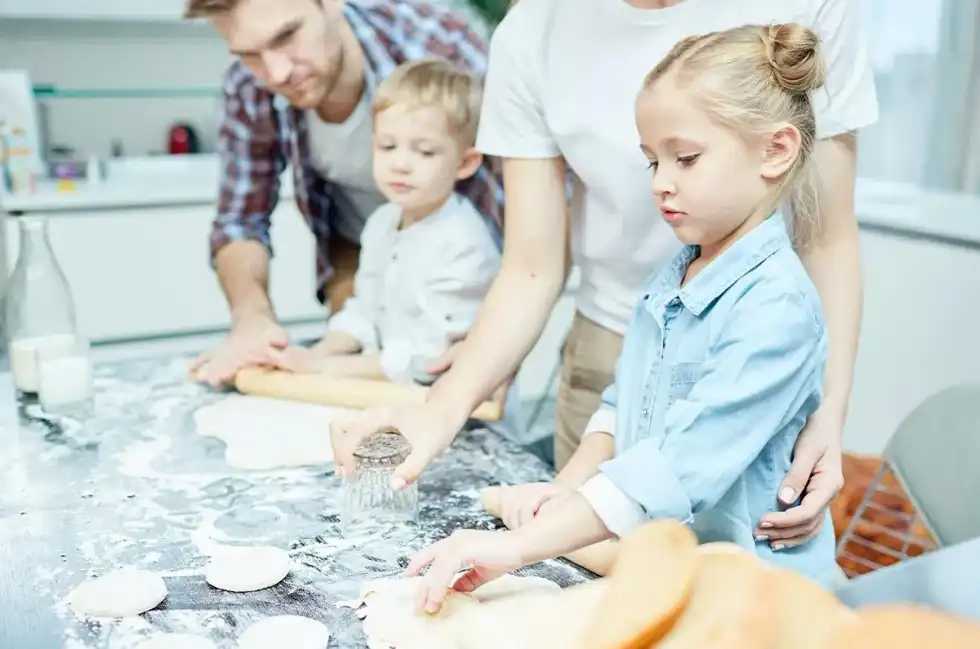 Little kids in the kitchen rolling out dough to making a dog shaped birthday cake with their parents helping.