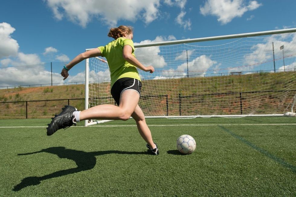 Low angle of female soccer player shooting the ball at the goal with a clear shadow on the turf.