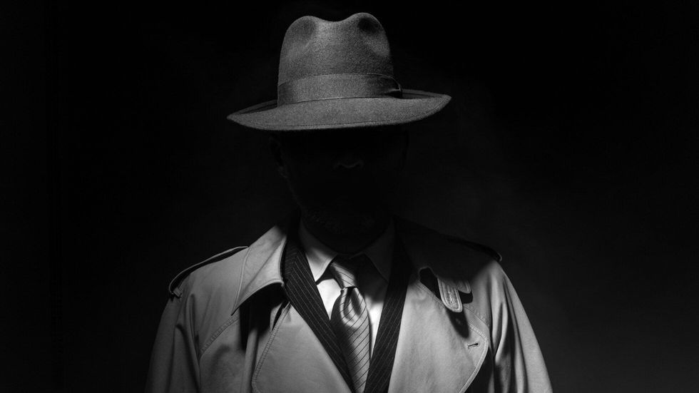 Man posing in the dark with a fedora hat and a trench coat like a Goodfella mafia member.
