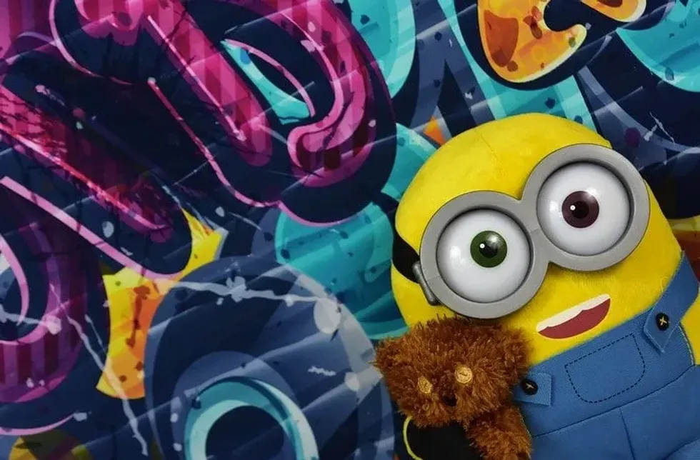 Minion standing in front of a graffiti wall holding a teddy bear.