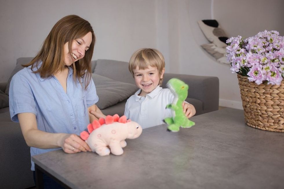 Mom and son have fun role-playing games with plush dinosaur toys on the table in the room. Dinosaurs move in their hands. Joyful communication and games with children, happy childhood, motherhood