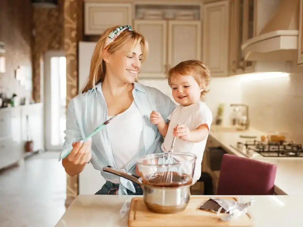 Mum in the kitchen holding her baby girl as she bakes a Barbie cake.