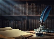 Old quill pen, books and vintage inkwell on wooden desk in the old office against the background of the bookcase and the rays of light.