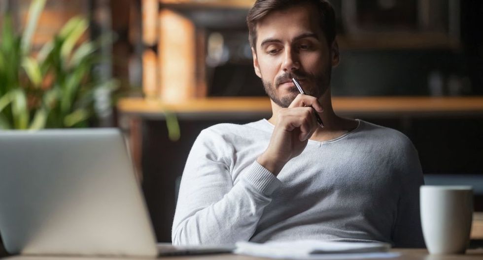 Pensive bearded man sitting at table drink coffee work at laptop thinking of problem solution