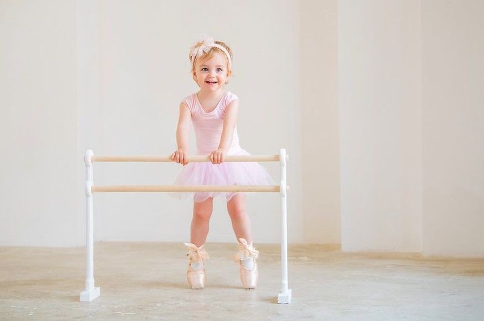 Portrait of cute blue-eyed baby ballerina in pink leotard and big pointe shoes standing near the ballet barre.