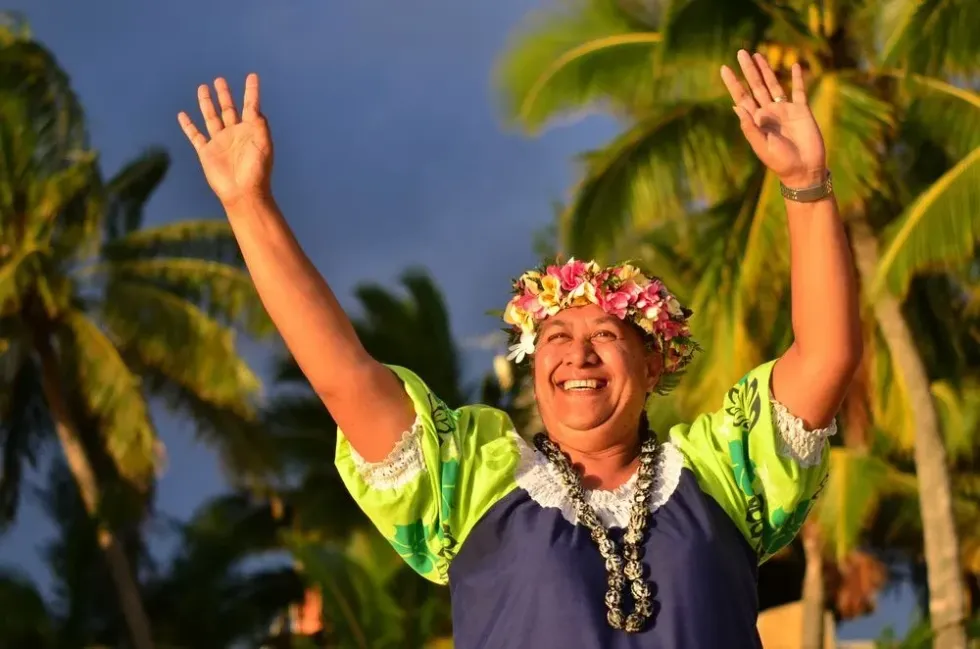 Portrait of happy active senior Polynesian Pacific Islander woman on tropical beach with palm trees in the background waving with her hands welcome hello