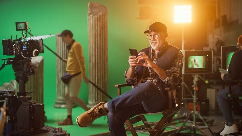 Prominent Successful Director Sitting in a Chair on a Break Using Smartphone. On the Studio Film Set with High-End Equipment Professional Crew Shooting High Budget Movie.