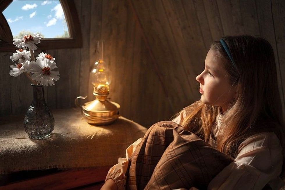 Sad girl sitting by the window in the attic with a kerosene lamp and paper flowers