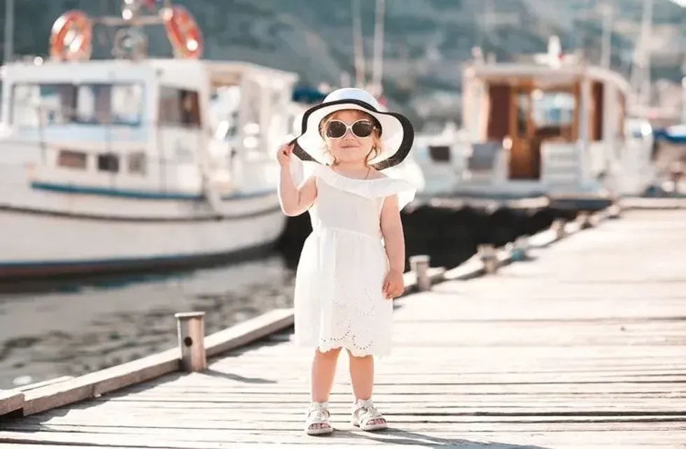 Smiling baby girl wearing white summer dress and hat walking on wooden pier at sea shore outdoors