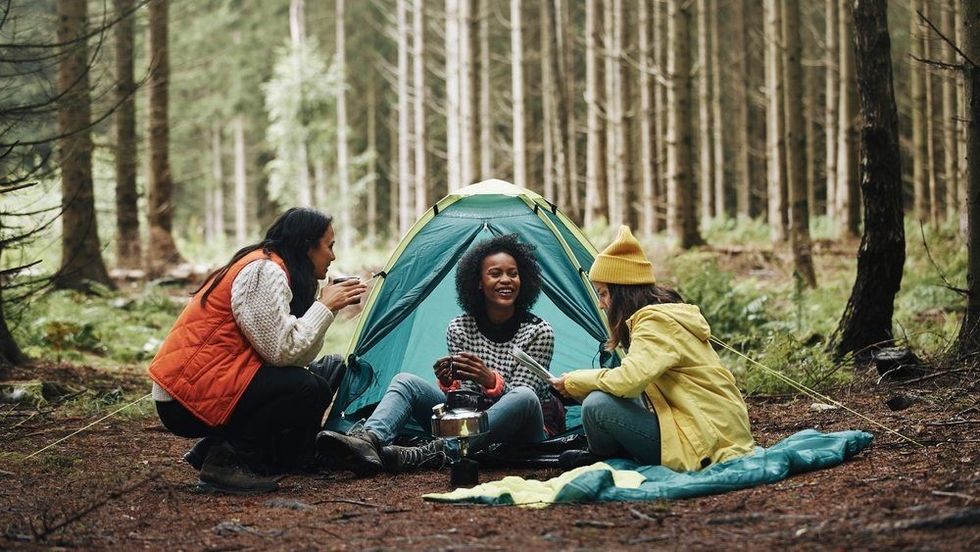 Smiling group of diverse young female friends sitting at their forest campsite drinking coffee and reading a trail map