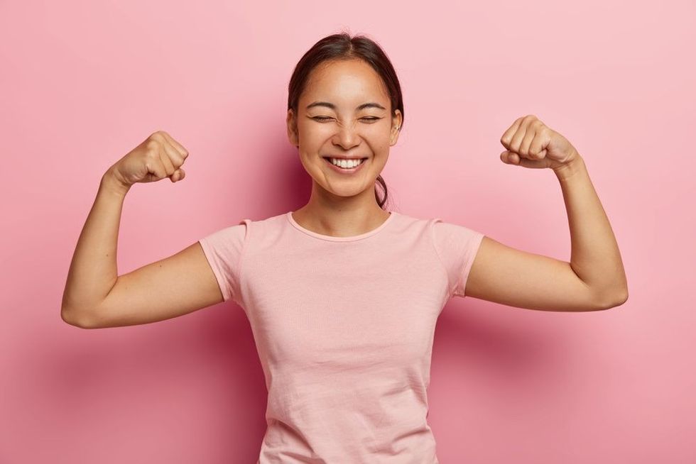 Strong woman with dark hair raises arms and shows biceps against pink background.