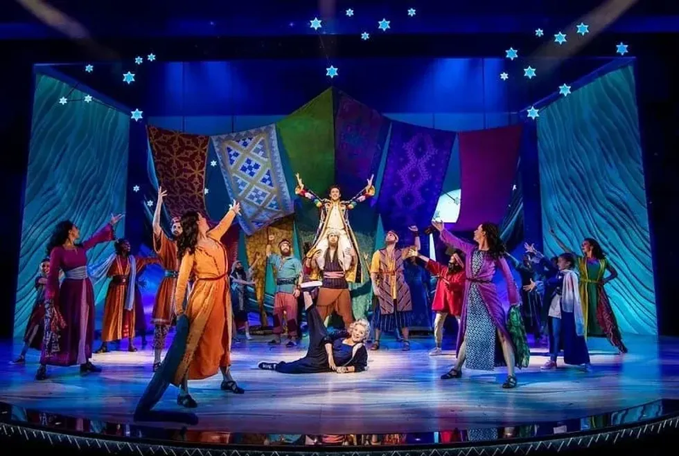 The cast of Joseph and the Amazing Technicolor Dreamcoat on stage pointing at the main character Joseph who is sitting on the shoulders of another actor. 