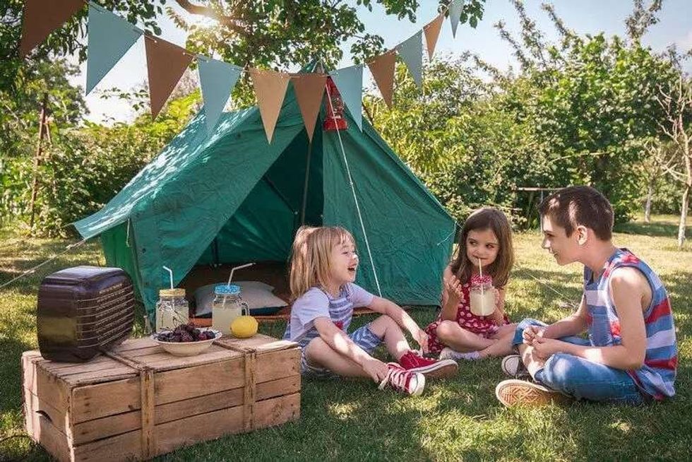 Three children in front of a green tent sitting on the grass.
