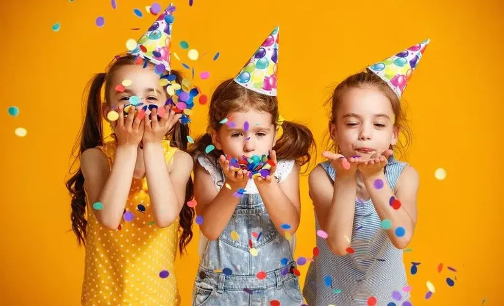 Three girls wearing party hats blowing confetti. 