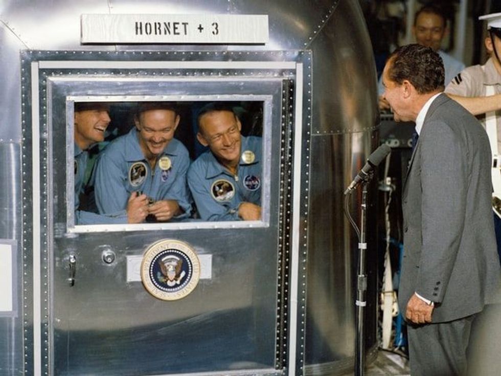 Three men laughing with a man in a suit speaking into a microphone toward them.