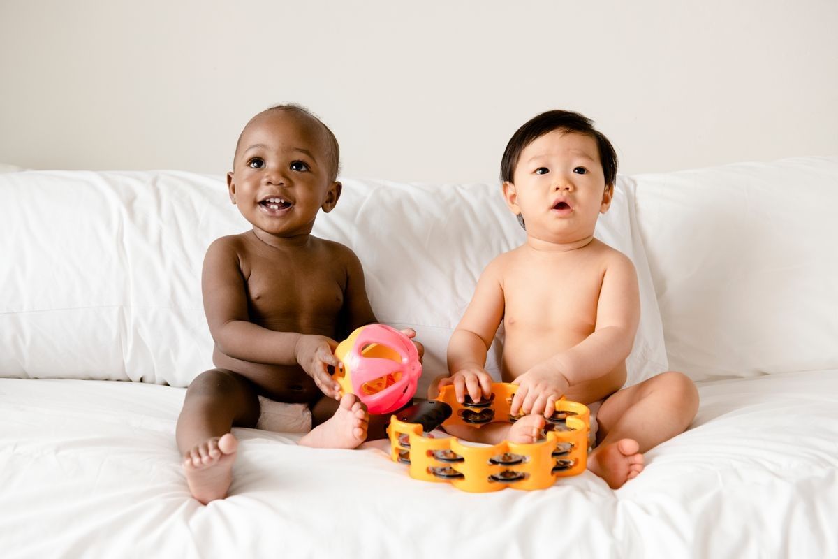 Two babies sitting on a bed, playing with musical toys, encapsulating the harmony of a melodic baby name.
