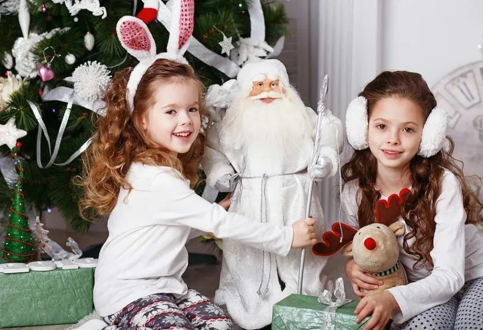 Two children are sitting surrounded by Christmas themed decorations and are smiling at the camera.