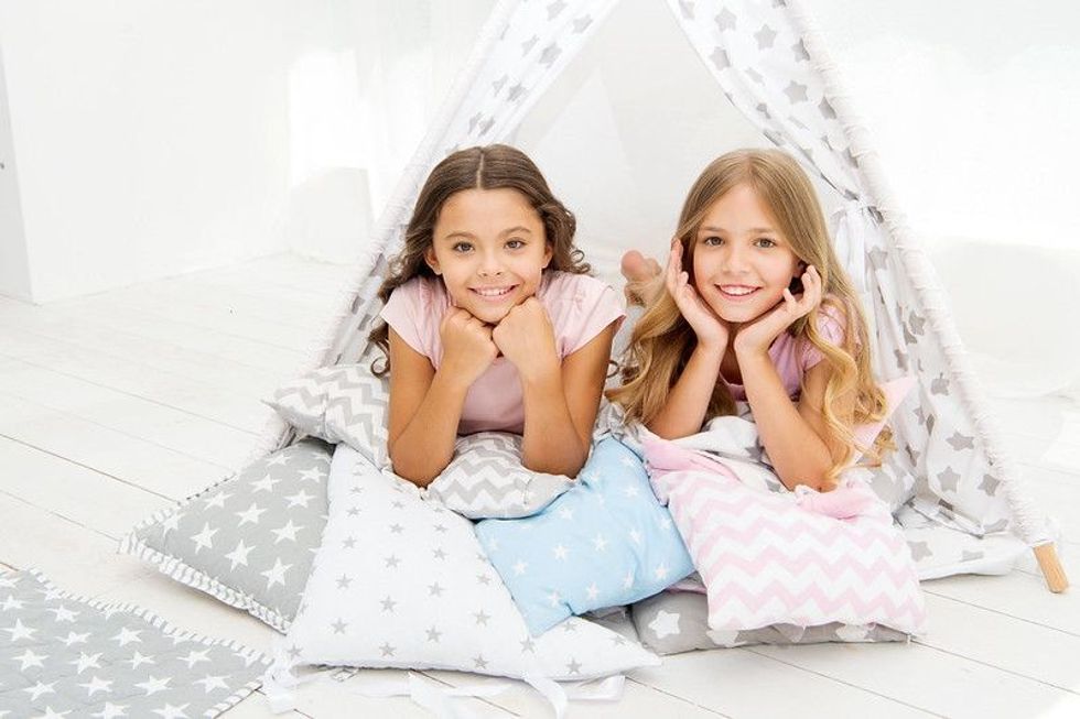 Two girls laying together and smiling, surrounded by bedding and pillows in a sleepover setting.