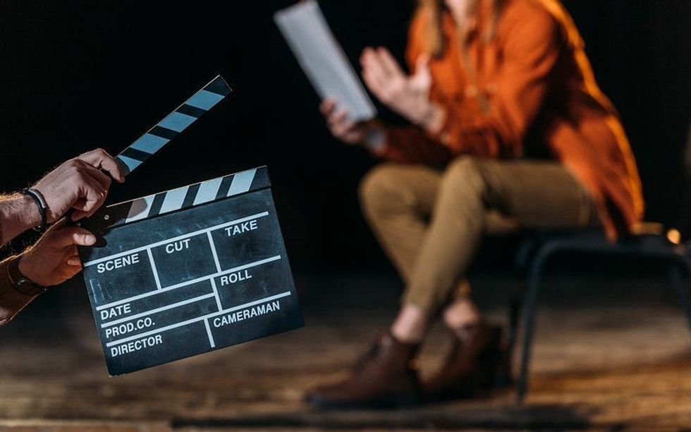 Two hands hold a clapperboard while someone is sitting on stage reading a book.