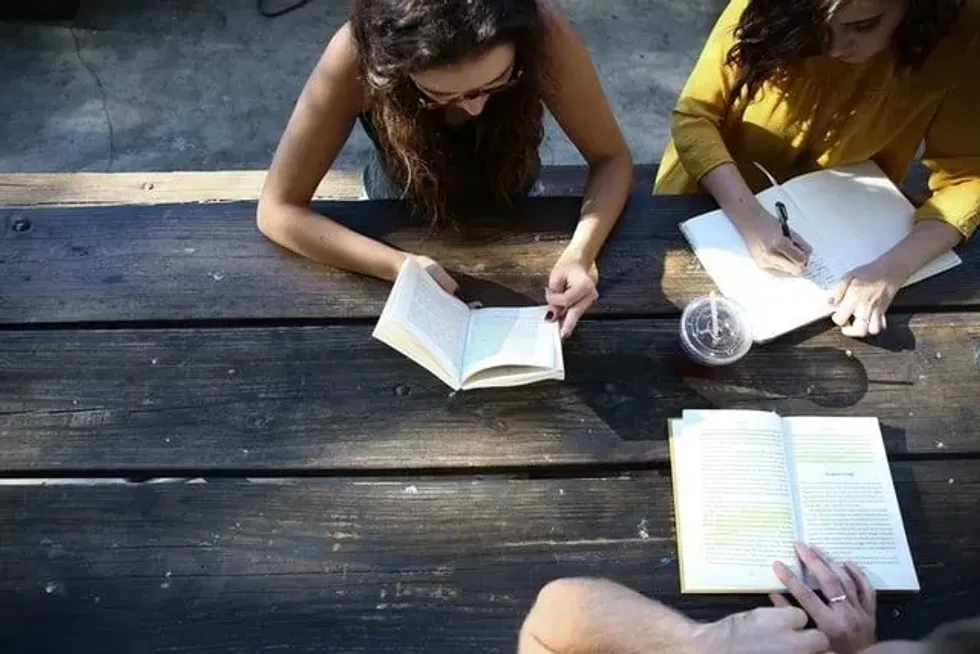 Two individuals engrossed in reading and writing at a rustic wooden table, with one reading a book and the other taking notes, possibly engaged in a game of modern literature trivia