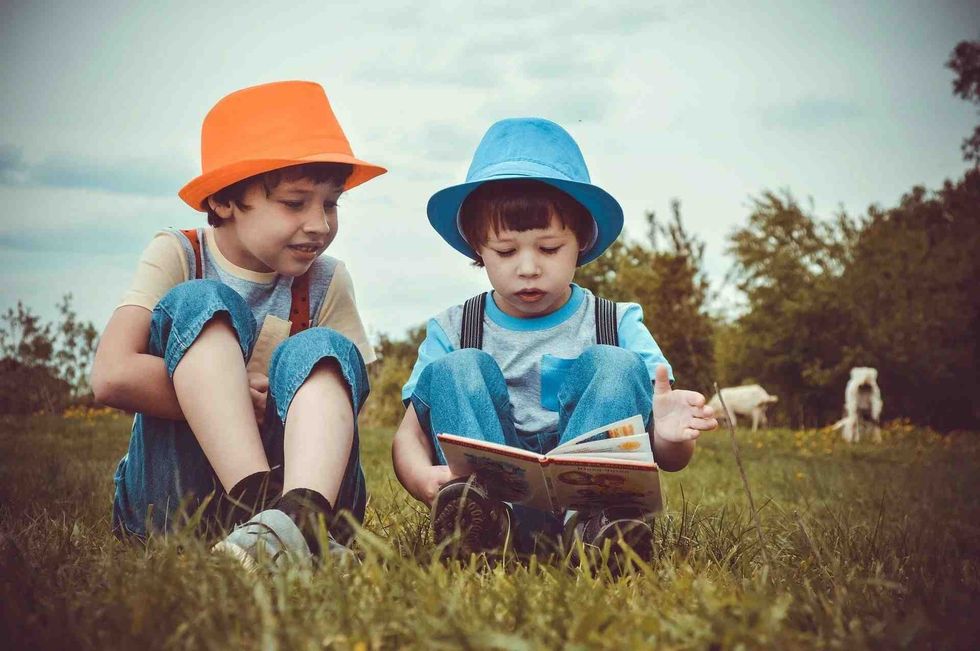 Two kids wearing hats are sitting in the meadows reading a book