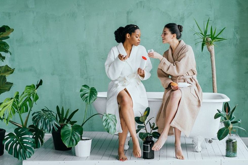Two women sitting on a bathtub are holding jars of cosmetics