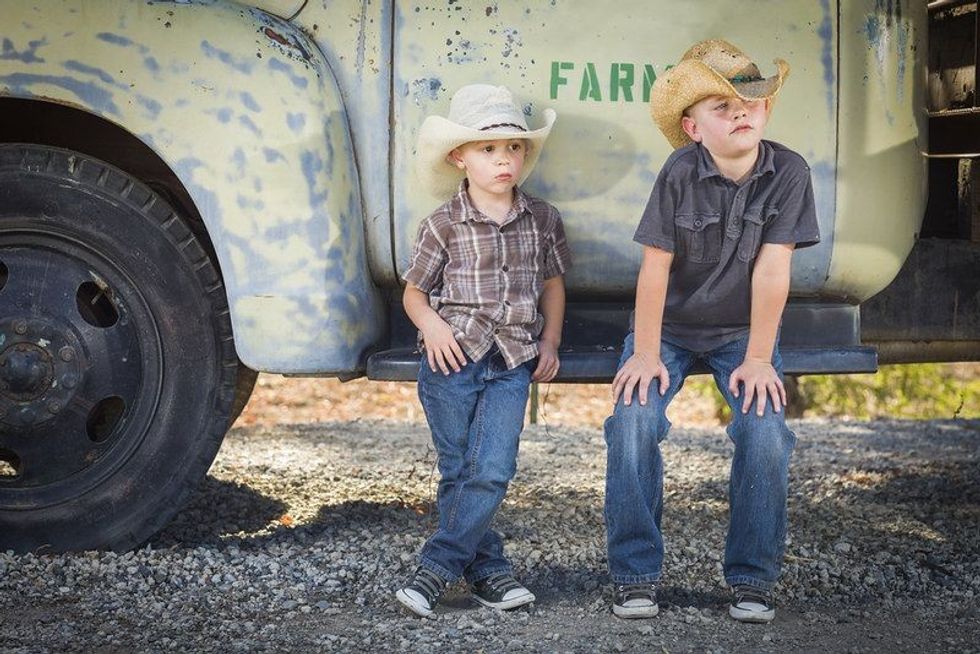 Two Young Boys Wearing Cowboy Hats Leaning Against an Antique Truck in a Rustic Country Setting.
