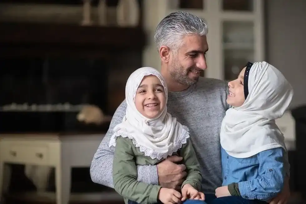 Two young girls wearing headscarves sat on their dad's lap happy and smiling.
