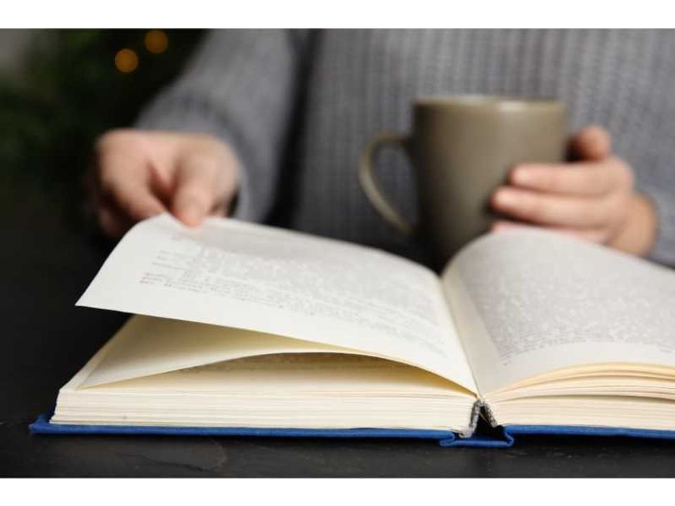 Woman with cup of beverage reading book at table