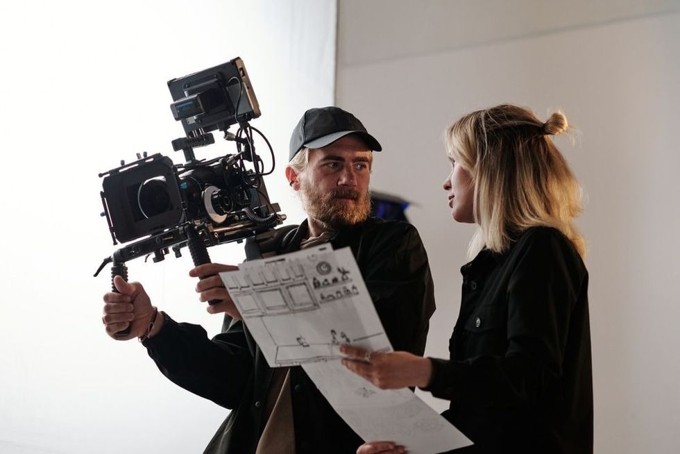 Young bearded cameraman and his assistant with documents consulting about shooting
