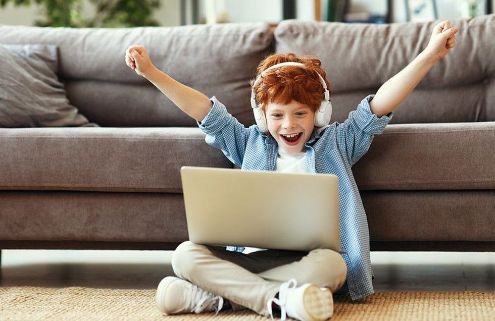 Young boy in headphones raising arms and smiling while sitting crossed legged on floor and celebrating victory in video game on laptop.