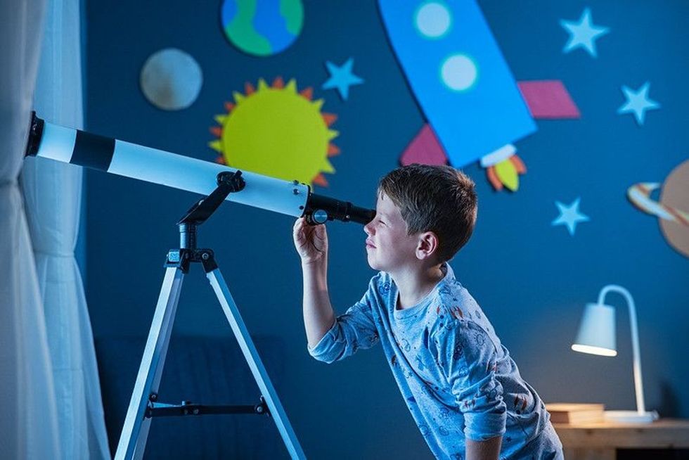 Young boy using telescope to see remote galaxy from room with decorated wall with rocket, planets, stars and spaceship.