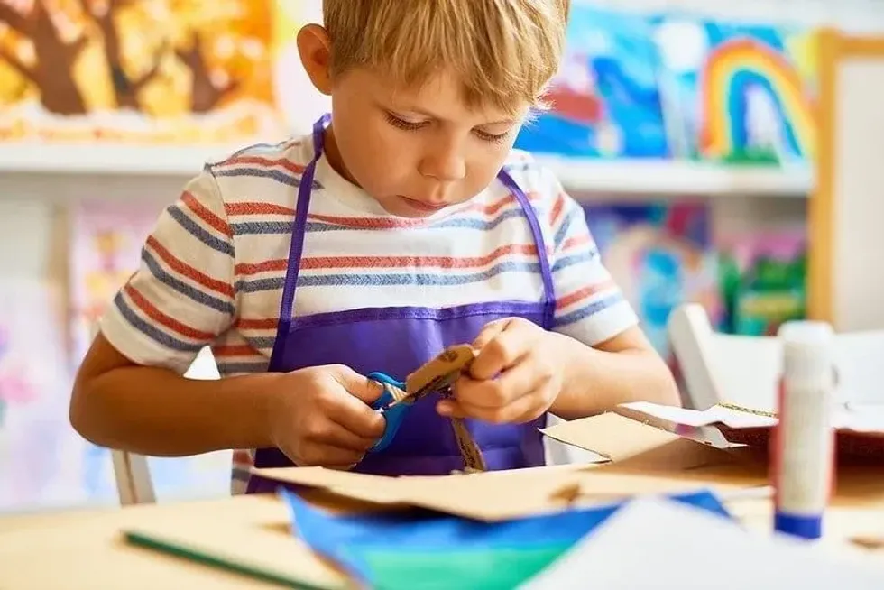 Young boy wearing an apron sat at the table cutting out cardboard.