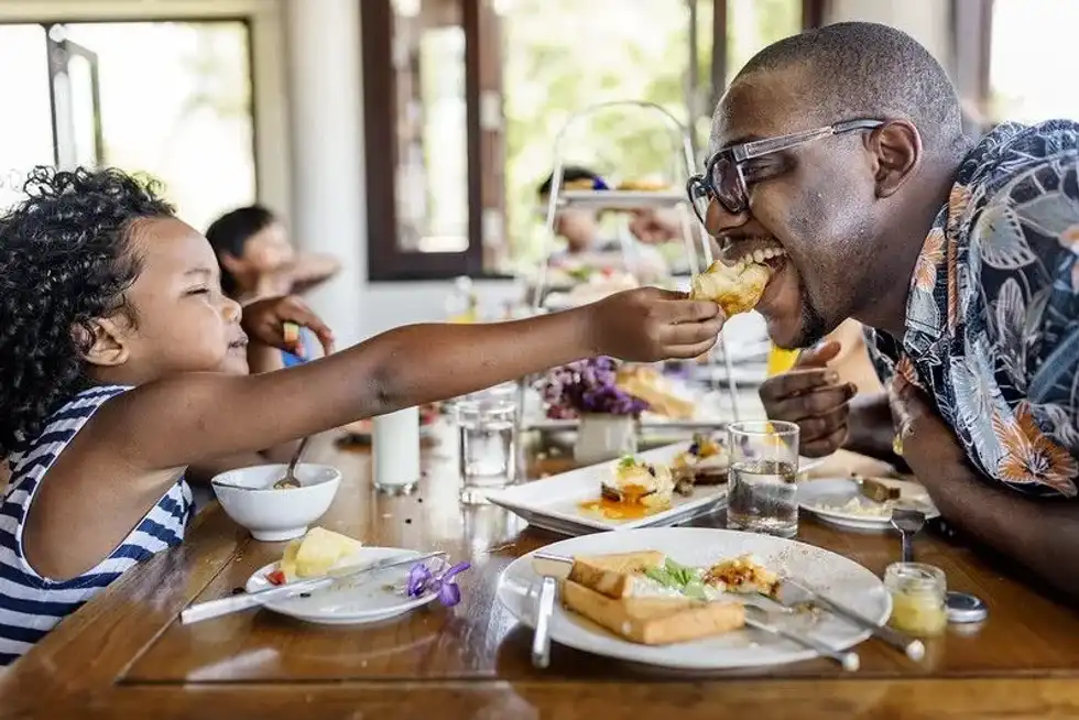 Young child attempting to playfully feed smiling man at the dinner table of a family restaurant.