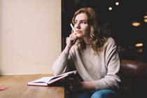 Young female novelist in baggy sweater thinking hand at chin and looking away through window while sitting at cafe table with open diary and pen in hand