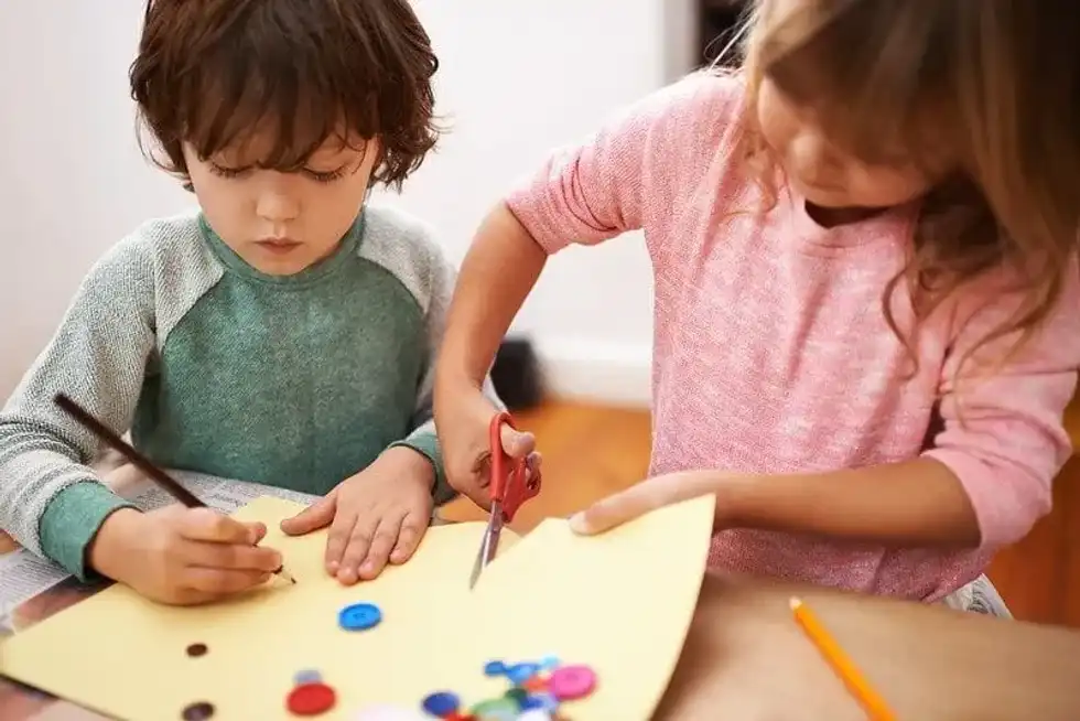 Young girl and boy sat at the table cutting paper and doing crafts at home.