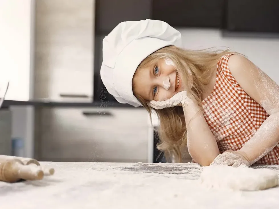 Young girl wearing a chef's hat smiling in the kitchen with flour on her face as she bakes a Scooby Doo cake.