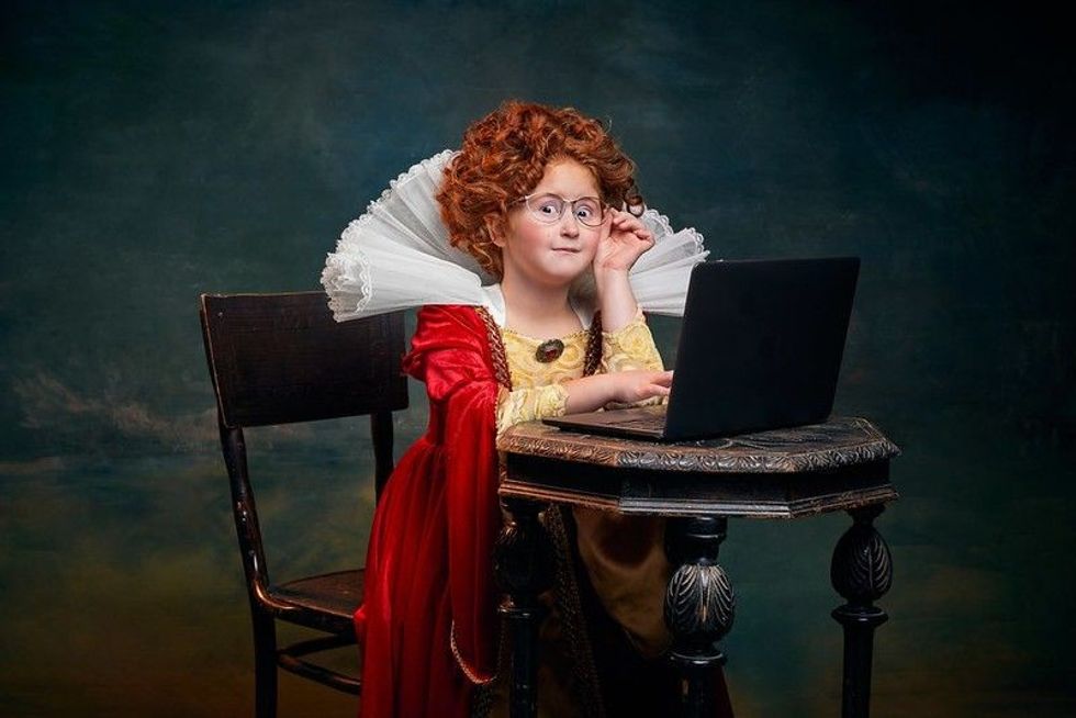 Young girl with curly red hair dressed in old fashioned royal person working on a laptop