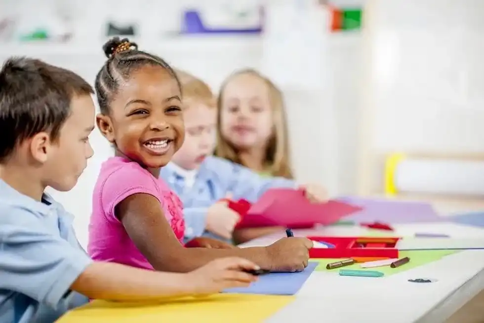 Young kids at the desk in a classroom, focusing on one girl who is smiling, cutting paper to make an origami unicorn.