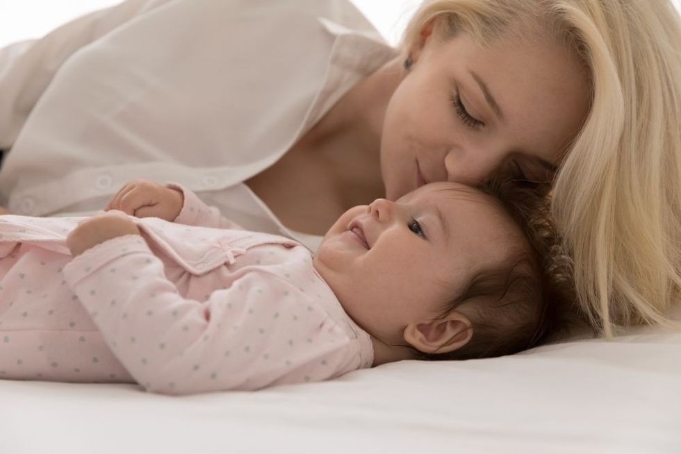 Young loving mother kisses on forehead her lovely babygirl, lying together on bed, close up cropped shot.