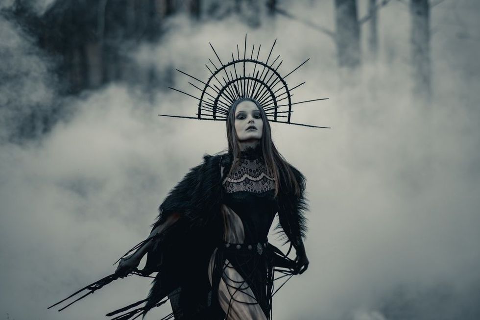 Young woman in image of witch stands in black dress and crown on her head in dark and foggy forest.